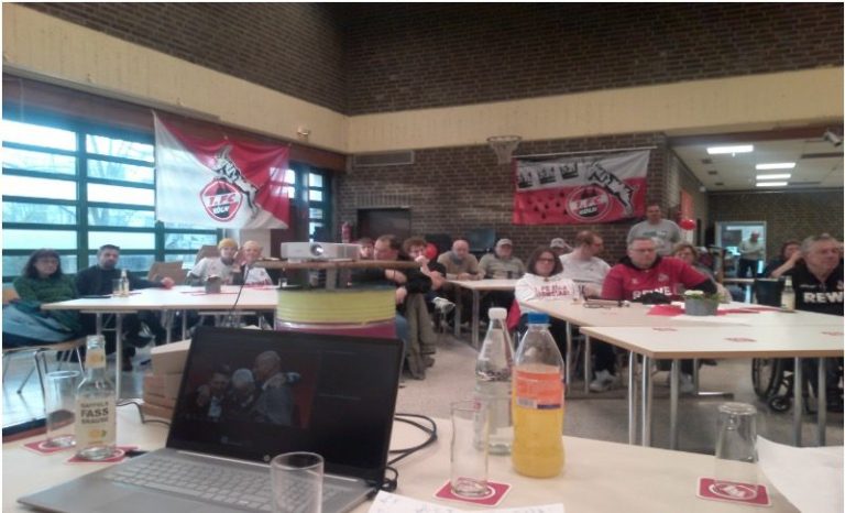 Gruppe von Menschen in einem Raum mit Tischen, Flaggen und Präsentationsmaterial.