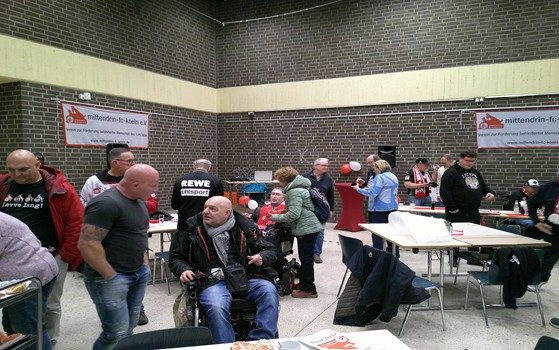 Menschen in einem Raum mit Tischen, einige stehen, andere sitzen, Banner im Hintergrund.
