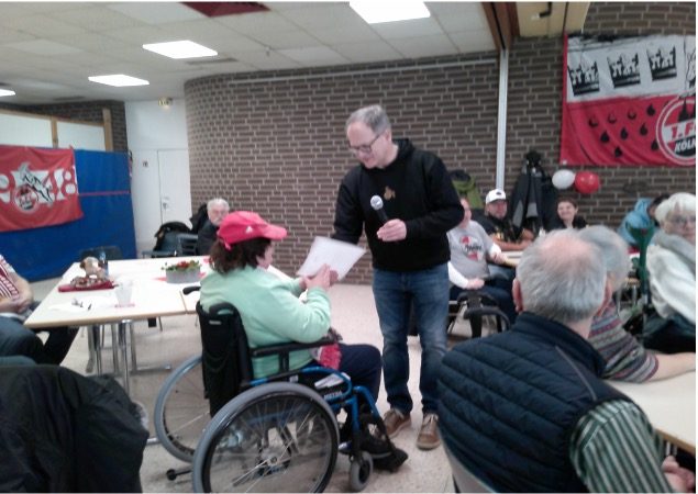 Eine Person im Rollstuhl erhält ein Dokument von einem Mann in einem Raum mit mehreren Personen.