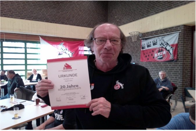 Mann hält ein Zertifikat in einem Raum mit Flaggen und Menschen im Hintergrund.