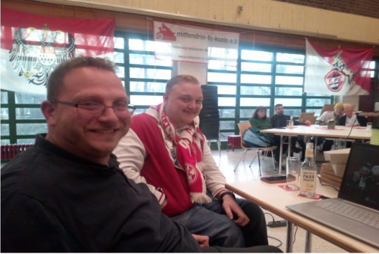 Zwei Männer lächeln, in einem Raum mit Tischen und sportlichen Dekorationen.